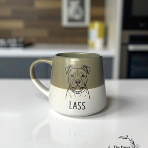 May include: A two-tone ceramic mug featuring a dog illustration and the name "LASS". The top half is olive green, the bottom half is white. The mug has a curved handle, suitable for hot drinks.