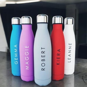 May include: Six personalised stainless steel water bottles with different names printed on them. The bottles are in various colours, including blue, purple, grey, red, and white.