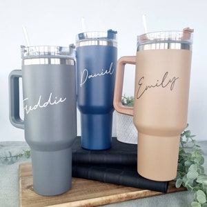 K&ouml;nnte beinhalten: Drei Edelstahl-Reisebecher mit Henkeln und Deckeln. Die Becher sind in Grau, Blau und Braun. Jeder Becher hat einen Namen: Freddie, Daniel und Emily.