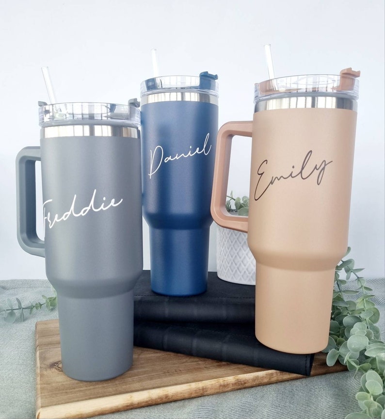 K&ouml;nnte beinhalten: Drei Edelstahl-Reisebecher mit Henkeln und Deckeln. Die Becher sind in Grau, Blau und Braun. Jeder Becher hat einen Namen: Freddie, Daniel und Emily.