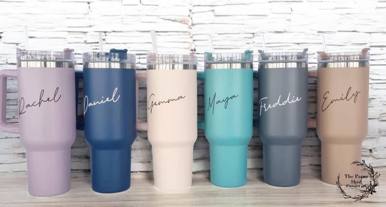 K&ouml;nnte beinhalten: Sechs personalisierte Edelstahl-Reisebecher mit Henkeln und Deckeln. Die Becher sind in verschiedenen Farben erh&auml;ltlich, darunter Lila, Blau, Beige, T&uuml;rkis, Grau und Braun. Jeder Becher tr&auml;gt einen Namen in schwarzer Schrift: Rachel, Daniel, Gemma, Maya, Freddie und Emily.