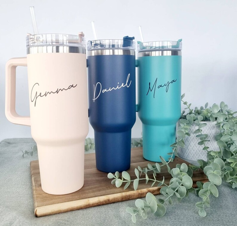 K&ouml;nnte beinhalten: Drei Edelstahl-Reisebecher mit Deckel und Strohhalmen. Die Becher sind mit den Namen "Gemma", "Daniel" und "Maya" personalisiert.