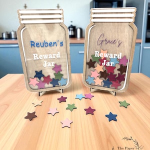 May include: Two wooden reward jars, each with a name and the words "Reward Jar." The jars are filled with colourful star-shaped tokens. Additional stars are scattered on the wooden surface. The Paper Shed Personalized Gifts logo is visible.