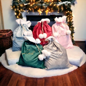 Può includere: Cinque sacchetti regalo di Natale personalizzati con pon pon. I sacchetti sono di diversi colori: azzurro, rosso, rosa, verde e grigio. I nomi "Emma", "Zach", "Rosie", "Jessie" e "Tuel" sono ricamati sui sacchetti con filo dorato.