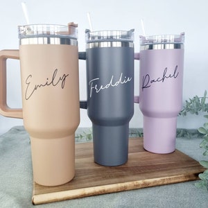 K&ouml;nnte beinhalten: Drei Edelstahl-Reisebecher mit Deckel und Henkeln. Die Becher sind in verschiedenen Farben: beige, grau und lila. Jeder Becher hat einen Namen darauf geschrieben: Emily, Freddie und Rachel.