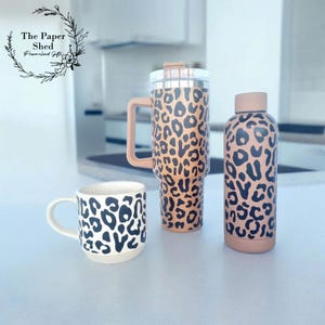 May include: Three stainless steel tumblers with a leopard print design. The tumblers are in different sizes, with the largest one having a handle. The tumblers are on a white surface.
