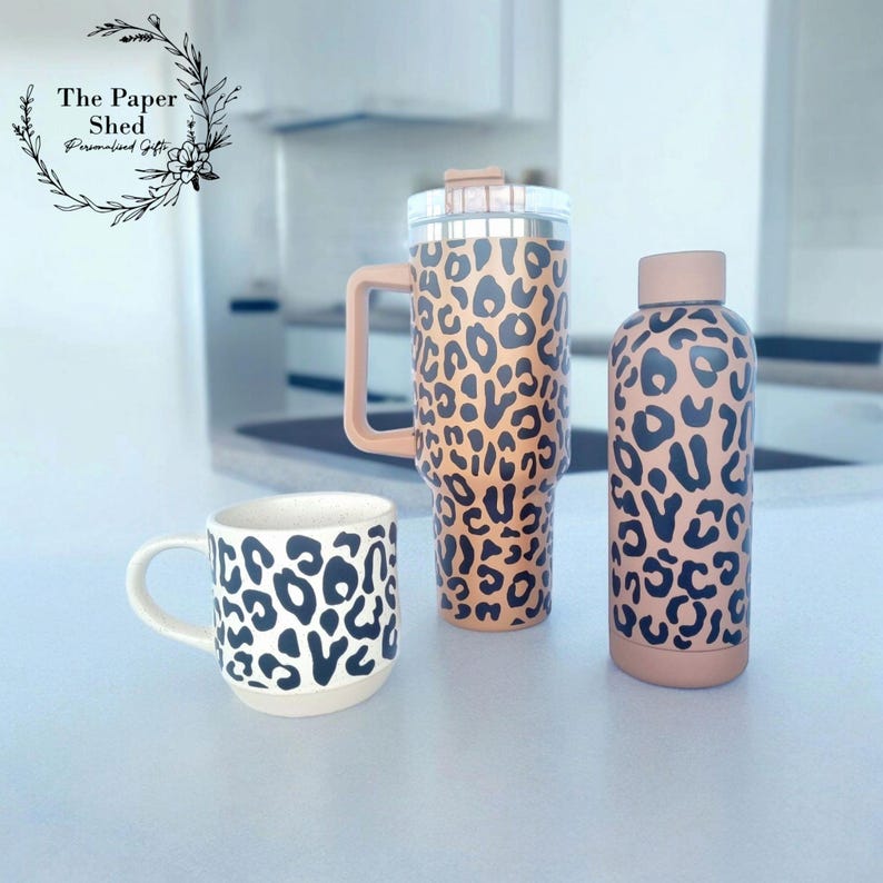 May include: Three stainless steel tumblers with a leopard print design. The tumblers are in different sizes, with the largest one having a handle. The tumblers are on a white surface.
