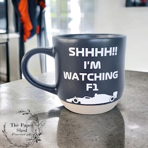May include: A dark grey ceramic mug with a white design that says "SHHHH!! I'M WATCHING F1" and a silhouette of a racing car.