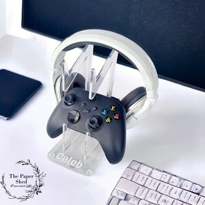 May include: A clear acrylic stand with the name "Caleb" engraved on the base. The stand holds a black video game controller and a white over-ear headset.