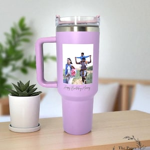 May include: A lilac insulated travel mug with a handle and a clear lid. The mug has a family photo and the text "Happy Birthday Mary". A small potted succulent sits on a wooden table next to the mug.