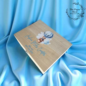 May include: A light brown wooden box with a teddy bear and hot air balloon design. The box is personalized with the name "Joshua Miles Carter", birth weight, and date in blue script. The background is a light blue fabric. The box is a personalized gift.