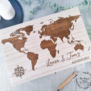 May include: A wooden box with a world map engraved on the lid. The map is in brown and the box is white. The text "Laura & Tom's Adventures" is engraved below the map. A compass is engraved in the bottom left corner of the box.
