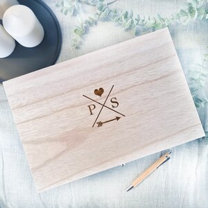 Può includere: Una scatola di legno con un'iscrizione personalizzata che presenta le iniziali "P" e "S" con un design a forma di cuore e freccia. La scatola è di colore marrone chiaro e ha una venatura del legno naturale.