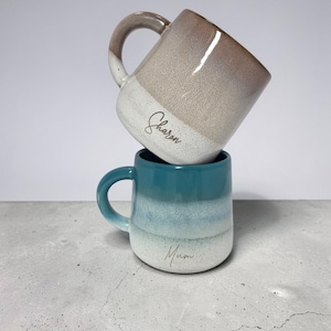May include: Two stacked ceramic mugs with a speckled glaze finish. The top mug is beige and white with the name Sharon written in script. The bottom mug is a teal and white gradient with the word Mum written in script on the lower section.