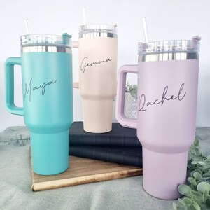 K&ouml;nnte beinhalten: Drei Edelstahl-Reisebecher mit Henkeln und Deckeln. Die Becher sind in verschiedenen Farben: T&uuml;rkis, Hellrosa und Beige. Jeder Becher hat einen Namen darauf geschrieben: Maya, Gemma und Rachel.