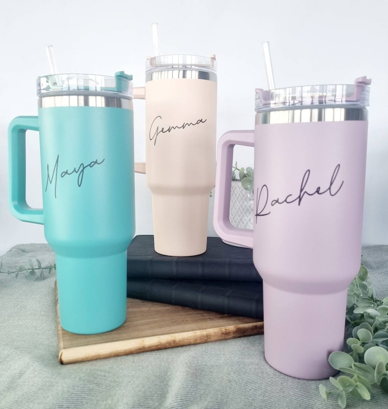 K&ouml;nnte beinhalten: Drei Edelstahl-Reisebecher mit Henkeln und Deckeln. Die Becher sind in verschiedenen Farben: T&uuml;rkis, Hellrosa und Beige. Jeder Becher hat einen Namen darauf geschrieben: Maya, Gemma und Rachel.