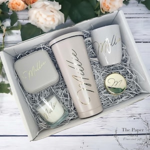 Può includere: Una scatola regalo bianca piena di articoli personalizzati, tra cui un bicchiere termico, una tazza, una candela e un piccolo cofanetto per gioielli. Tutti gli articoli sono decorati con il nome "Millie" in lettere dorate.