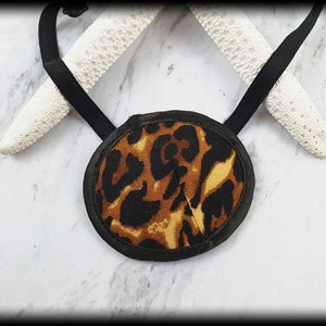 Puede incluir: Un parche en el ojo con un borde negro y un diseño de estampado de leopardo en marrón y negro. El parche en el ojo tiene una correa elástica negra para asegurarlo. El fondo incluye dos objetos blancos texturizados.