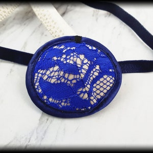 以下が含まれることがあります： ネイビーブルーのサテンの縁とストラップが付いた青いアイパッチ。中央には、鮮やかな青いレースが重ねられた繊細なクリーム色のレースのデザインが施されています。アイパッチは快適さとスタイルを考慮して設計されています。