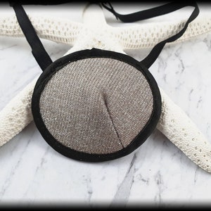 Pode incluir: Uma tapa-olho prateada com uma borda preta e tiras elásticas pretas. A tapa-olho é redonda e tem uma superfície texturizada. O fundo inclui uma estrela do mar branca e uma superfície de mármore.