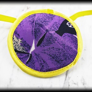 Pode incluir: Uma tapa-olho redonda com um padrão floral roxo e uma borda amarela. A tapa-olho tem uma alça amarela para prendê-la. O tecido tem uma aparência acolchoada.