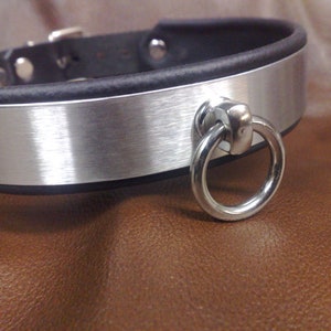 Könnte beinhalten: Ein silbernes Metallhalsband mit schwarzem Lederuntergrund und einem daran befestigten silbernen Ring.