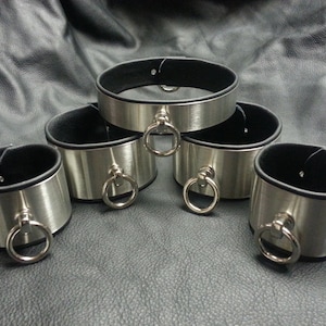 Puede incluir: Cinco brazaletes de metal plateado con forro de cuero negro. Cada brazalete tiene un anillo plateado.