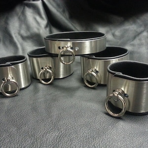 Puede incluir: Cinco brazaletes de metal plateado con forro de cuero negro y un anillo plateado en cada brazalete.