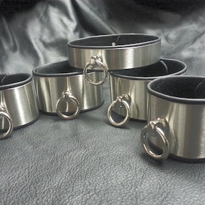 Puede incluir: Cinco brazaletes de metal plateado con ribete de cuero negro y un anillo plateado en cada brazalete.
