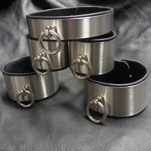 Puede incluir: Cuatro brazaletes de metal plateado con forro de cuero negro y anillos plateados. Los brazaletes est&aacute;n apilados uno encima del otro.