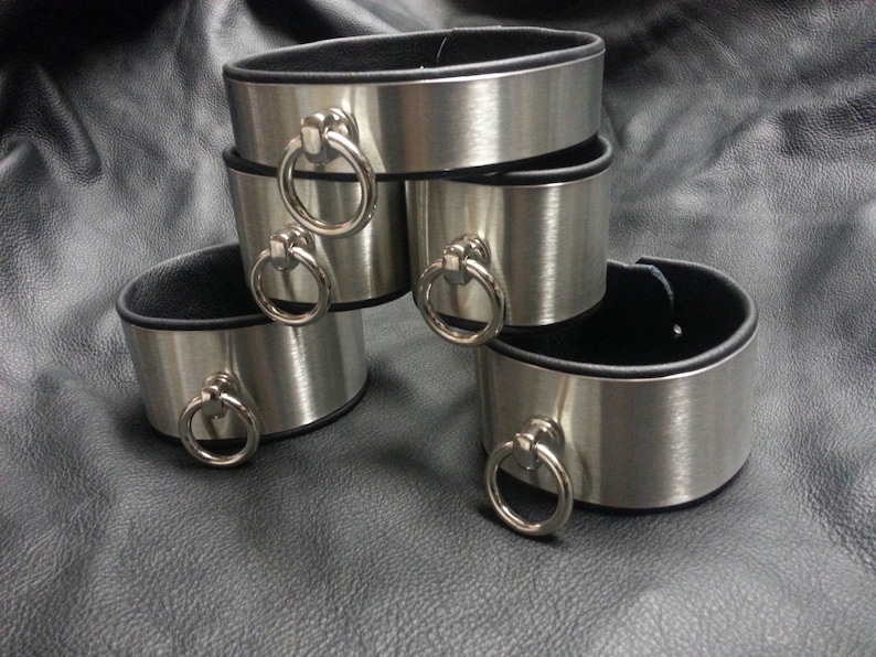 Puede incluir: Cuatro brazaletes de metal plateado con forro de cuero negro y anillos plateados. Los brazaletes est&aacute;n apilados uno encima del otro.