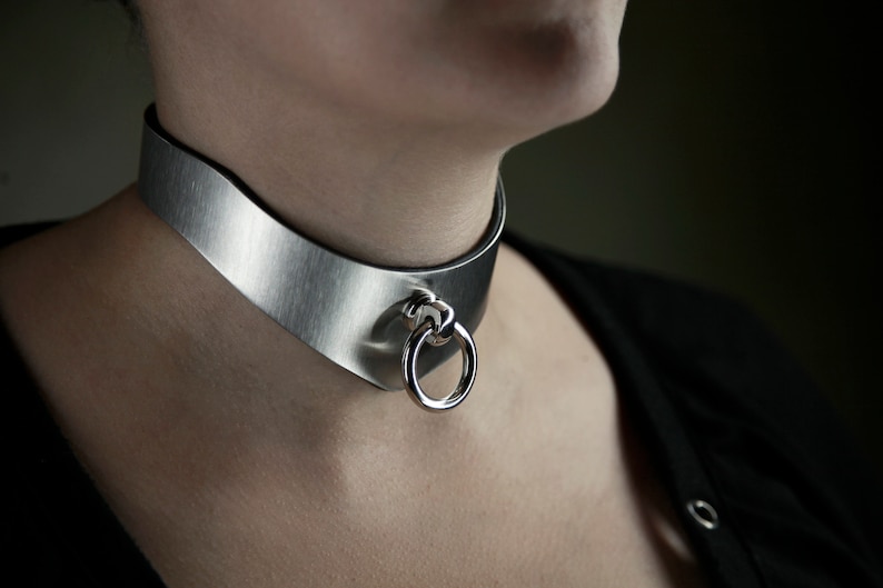 May include: A silver metal choker necklace with a smooth, brushed finish. The necklace has a large, round, silver ring attached to it.