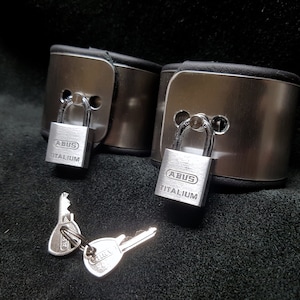 Puede incluir: Dos esposas de metal plateado con correas de cuero negro y candados plateados. Los candados est&aacute;n etiquetados con "ABUS TITANIUM". Se incluyen dos llaves plateadas.