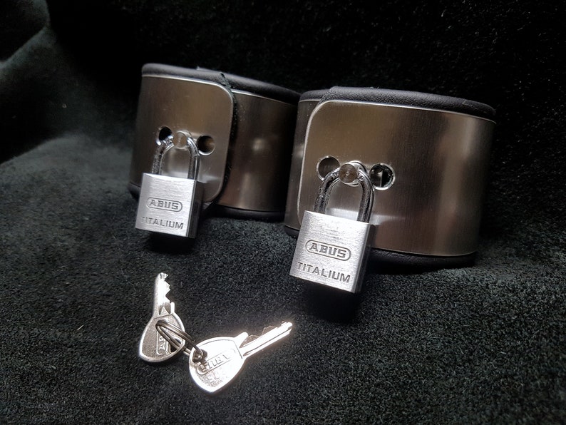 Puede incluir: Dos esposas de metal plateado con correas de cuero negro y candados plateados. Los candados est&aacute;n etiquetados con "ABUS TITANIUM". Se incluyen dos llaves plateadas.