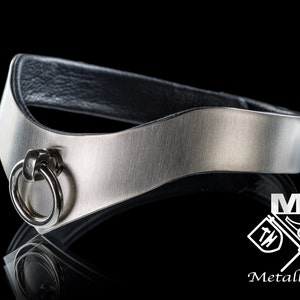 May include: A silver metal collar with a black leather backing and a silver ring. The collar is curved and has a brushed finish. The ring is attached to the collar with a small silver loop. The collar is made by Metall Geyer.