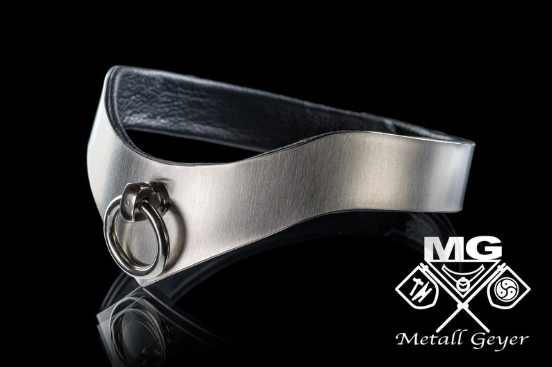 May include: A silver metal collar with a black leather backing and a silver ring. The collar is curved and has a brushed finish. The ring is attached to the collar with a small silver loop. The collar is made by Metall Geyer.