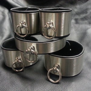 Puede incluir: Cuatro brazaletes de metal plateado con forro de cuero negro y un anillo plateado en cada brazalete.
