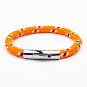 May include: A braided orange rope bracelet featuring small, repeating white and dark blue diagonal accents throughout the cord. The bracelet is secured with a polished, cylindrical silver-toned metal clasp that connects the two ends of the rope.
