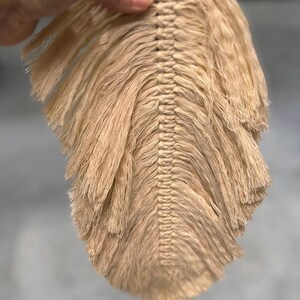 May include: A beige macrame feather with a central woven spine and fringed edges. The handmade wall decor is held up by a hand. The feather design is a neutral color, suitable for bohemian or minimalist decor.