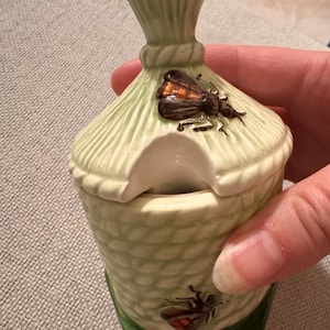 Op de afbeelding: Een lichtgroene keramische honingpot met een gestructureerd bijenkorfpatroon en een bijpassend deksel. De pot heeft een kleine uitsparing voor een lepel en is versierd met geschilderde bruine en oranje bijen op het deksel en de pot.