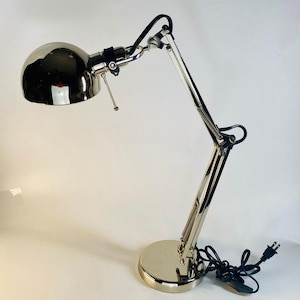 May include: A silver desk lamp with a flexible arm and a round, silver shade. The lamp has a black cord and plug.