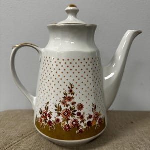 May include: A white porcelain teapot featuring a delicate pattern of small brown polka dots and a cluster of orange and brown floral motifs near the base. The teapot includes a matching lid with a gold-toned finial and gold trim on the handle.