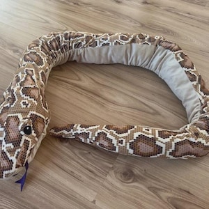 May include: A plush snake toy in shades of brown, tan, and white, with a patterned design. The snake has a light gray underside and a blue tongue. The toy is lying on a wooden surface.