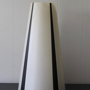 May include: A tall, off-white lamp shade with a conical shape. Two vertical black stripes run along the sides of the shade. The lamp shade is on a dark surface against a white wall.