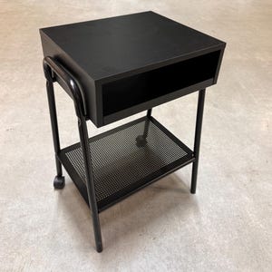 May include: Black rolling side table with a rectangular top and a lower shelf. The table has a drawer-like opening and a perforated metal shelf. The frame is made of black metal and has wheels for easy movement.