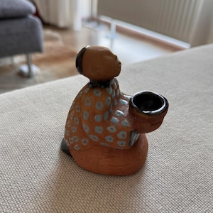 Puede incluir: Figura de cerámica de una persona arrodillada sosteniendo un cuenco pequeño. La figura tiene una cabeza marrón y viste una prenda naranja con lunares azules. El cuenco es negro y la figura está sobre una superficie de color neutro.