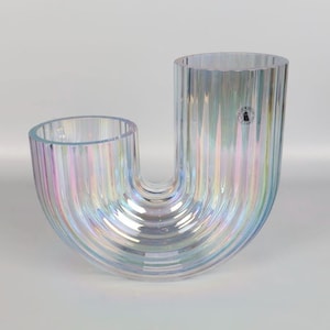 May include: A U-shaped glass vase with a vertical ribbed texture and an iridescent finish that reflects light in shades of pink, blue, and silver. The vase features a small circular sticker with a graphic icon on the right side of the rim.