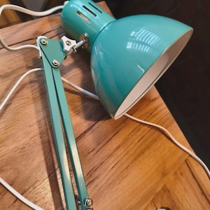 May include: A turquoise desk lamp with an adjustable metal arm, a wide dome-shaped shade, and a white power cord. The lamp features a white plastic adjustment knob at the joint and a separate white clamp mechanism for mounting to a flat surface.