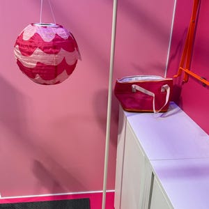 May include: A pink and white paper lantern hangs from a white metal stand with a wavy top. A pink insulated tote bag sits on a white cabinet. The background is a solid pink wall.
