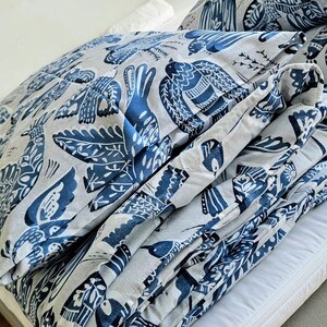 Può includere: Un set di copripiumino e cuscino piegato con un motivo ripetuto di uccelli blu stilizzati e fogliame su sfondo grigio chiaro. La biancheria da letto è ordinatamente impilata, mostrando il design dettagliato e il contrasto cromatico.